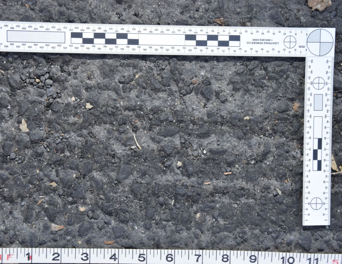 Close view of uneven asphalt with small stones and plant fragments, framed by an L-shaped scale and measuring tape.