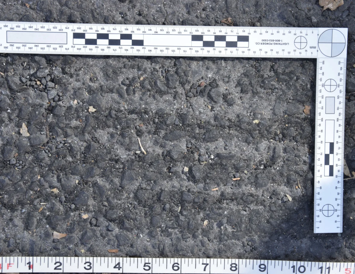 Close view of uneven asphalt with small stones and plant fragments, framed by an L-shaped scale and measuring tape.