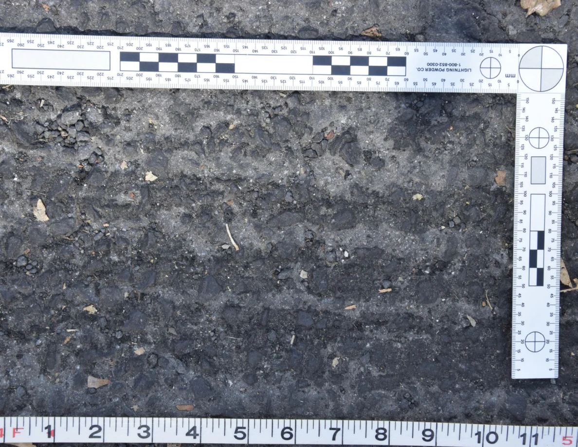 Close view of uneven asphalt with small stones and plant fragments, framed by an L-shaped scale and measuring tape.