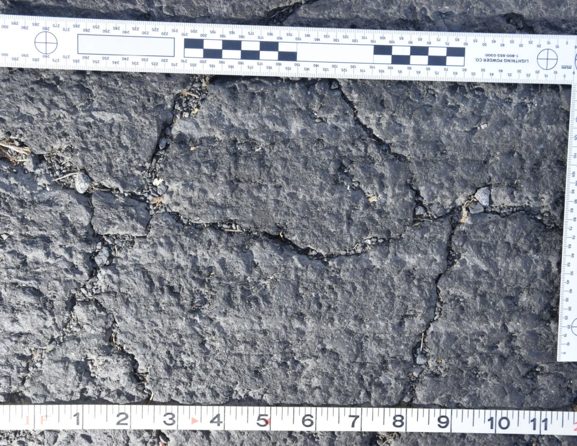 Intersecting cracks in asphalt with small pebbles lodged in the gaps, beside an L-shaped scale and measuring tape.