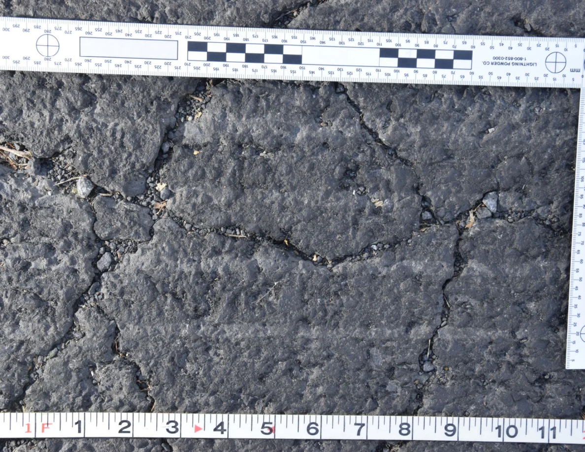 Intersecting cracks in asphalt with small pebbles lodged in the gaps, beside an L-shaped scale and measuring tape.