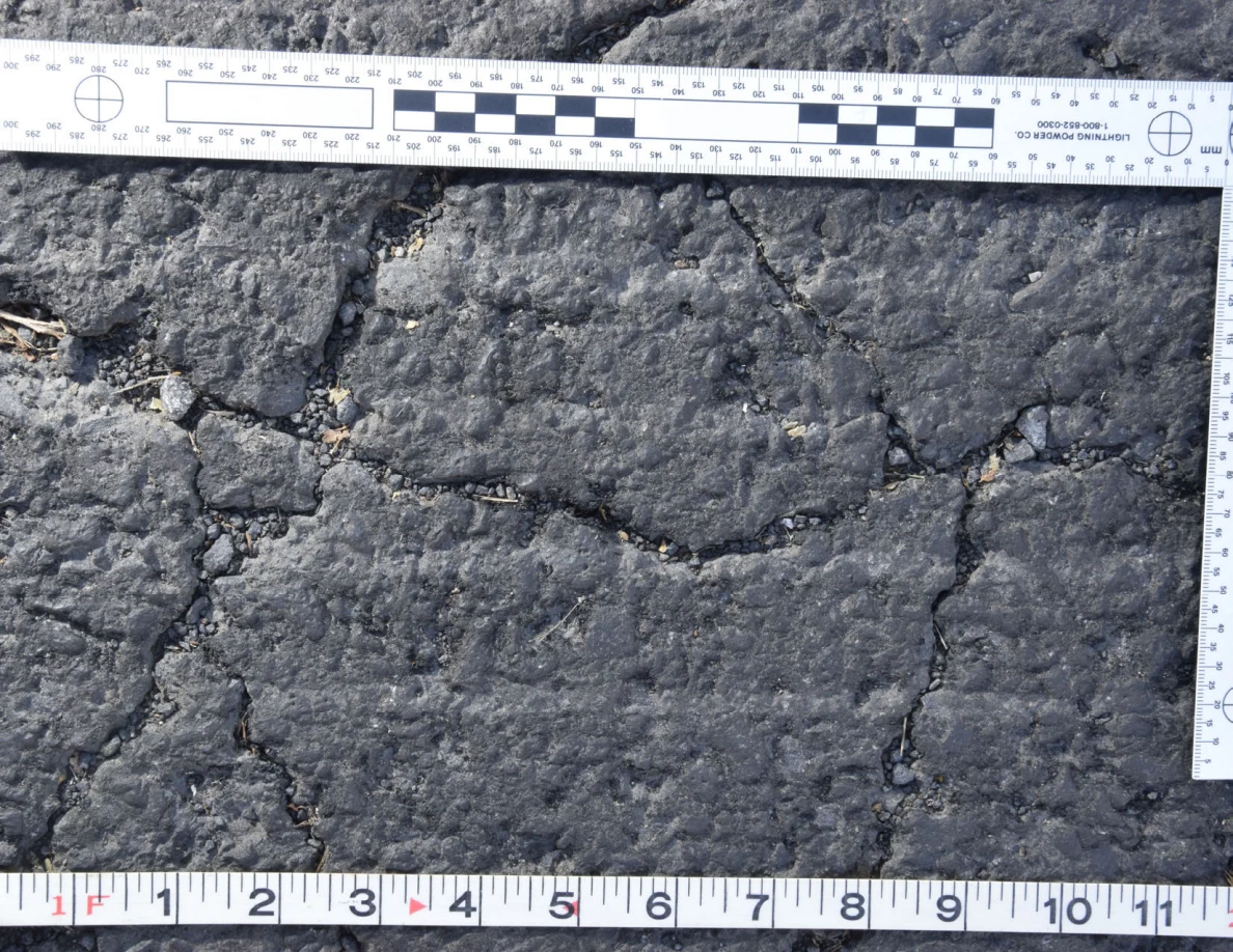 Intersecting cracks in asphalt with small pebbles lodged in the gaps, beside an L-shaped scale and measuring tape.