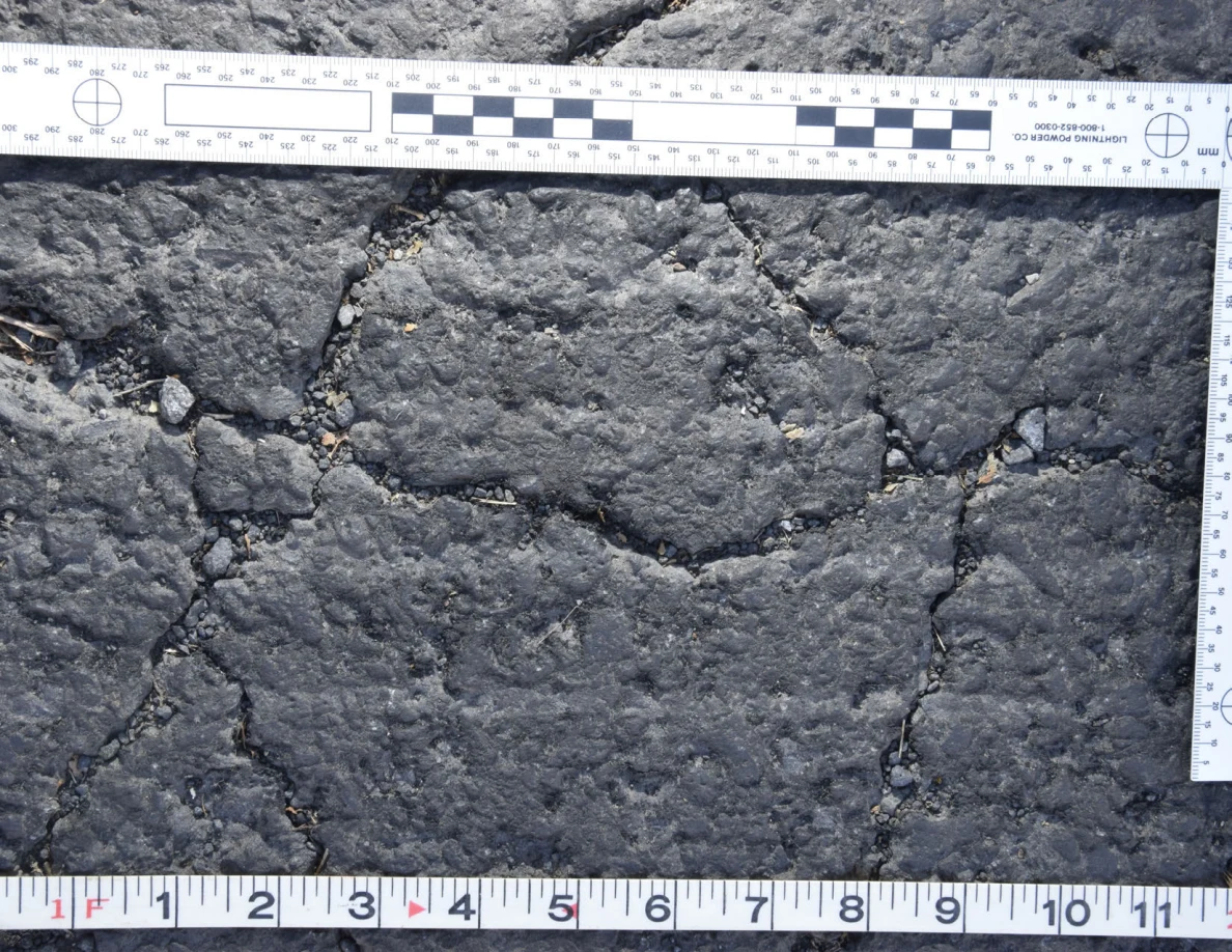 Intersecting cracks in asphalt with small pebbles lodged in the gaps, beside an L-shaped scale and measuring tape.