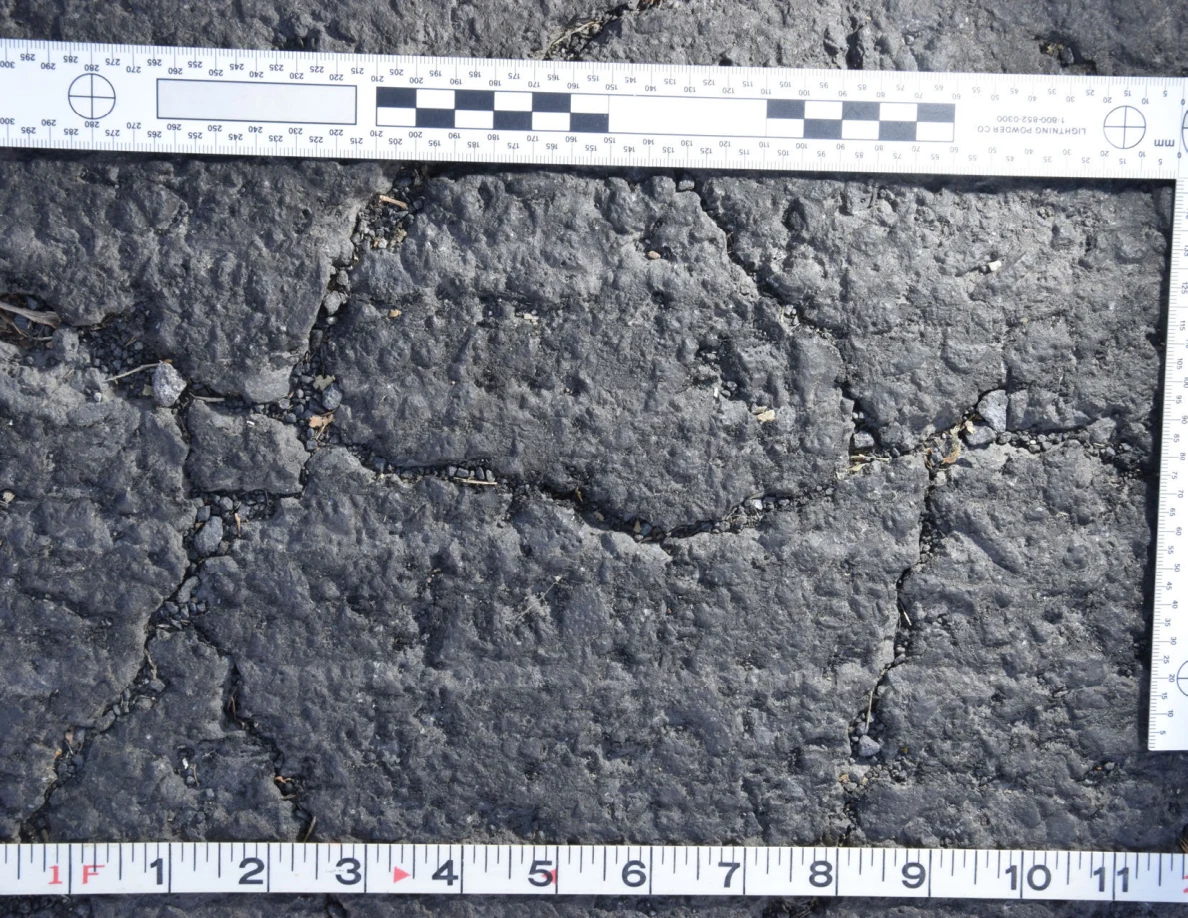 Intersecting cracks in asphalt with small pebbles lodged in the gaps, beside an L-shaped scale and measuring tape.