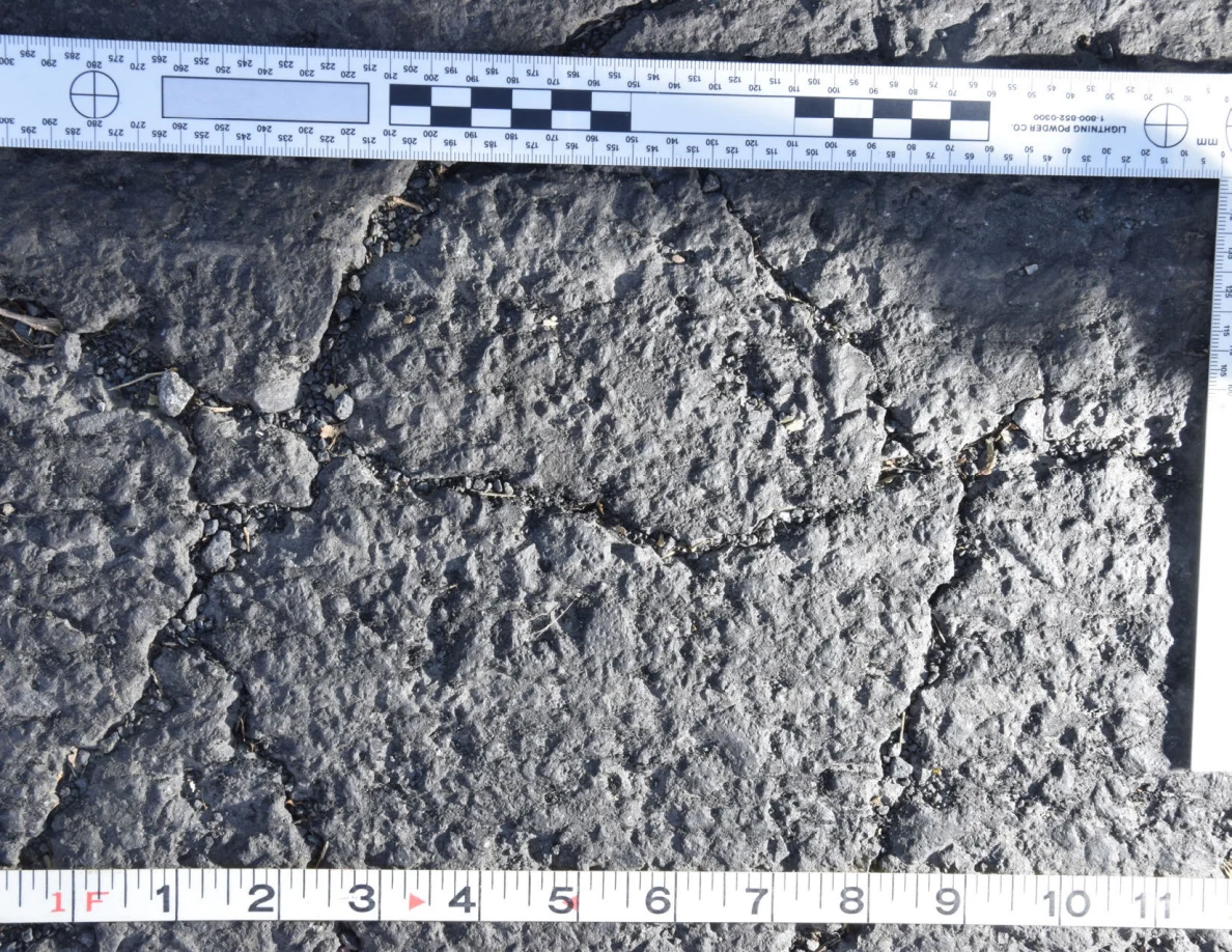 Intersecting cracks in asphalt with small pebbles lodged in the gaps, beside an L-shaped scale and measuring tape.