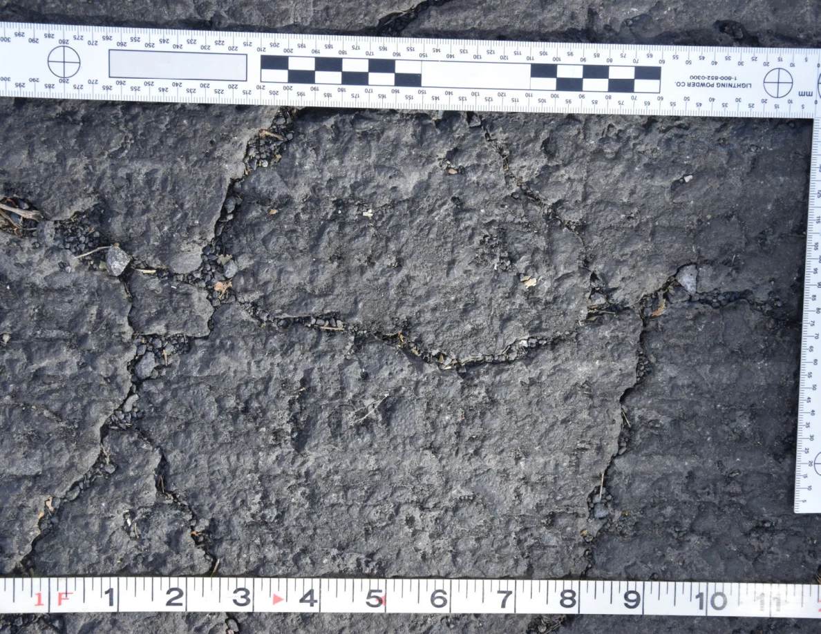 Intersecting cracks in asphalt with small pebbles lodged in the gaps, beside an L-shaped scale and measuring tape.