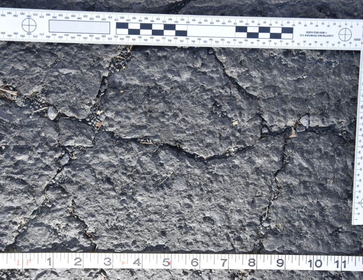 Intersecting cracks in asphalt with small pebbles lodged in the gaps, beside an L-shaped scale and measuring tape.