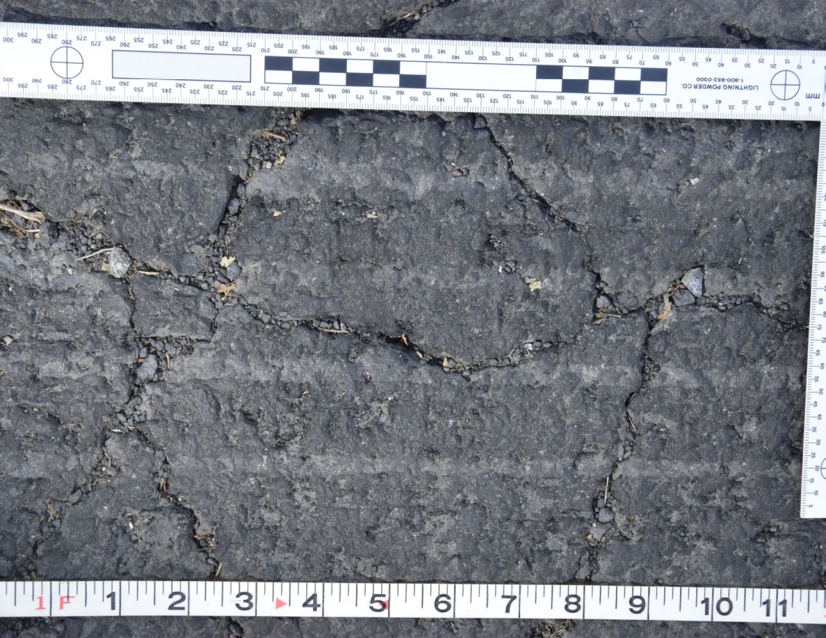 Intersecting cracks in asphalt with small pebbles lodged in the gaps, beside an L-shaped scale and measuring tape.