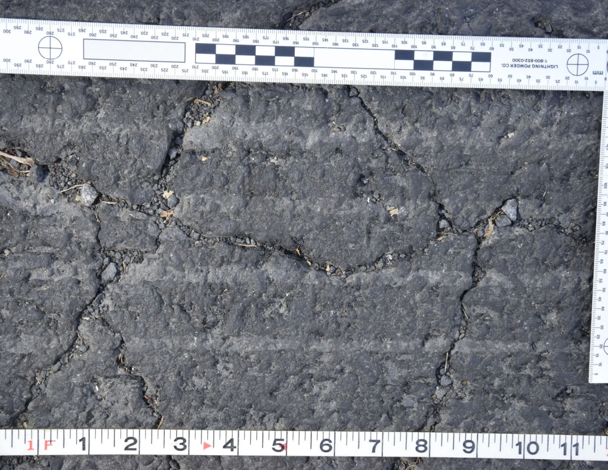 Intersecting cracks in asphalt with small pebbles lodged in the gaps, beside an L-shaped scale and measuring tape.