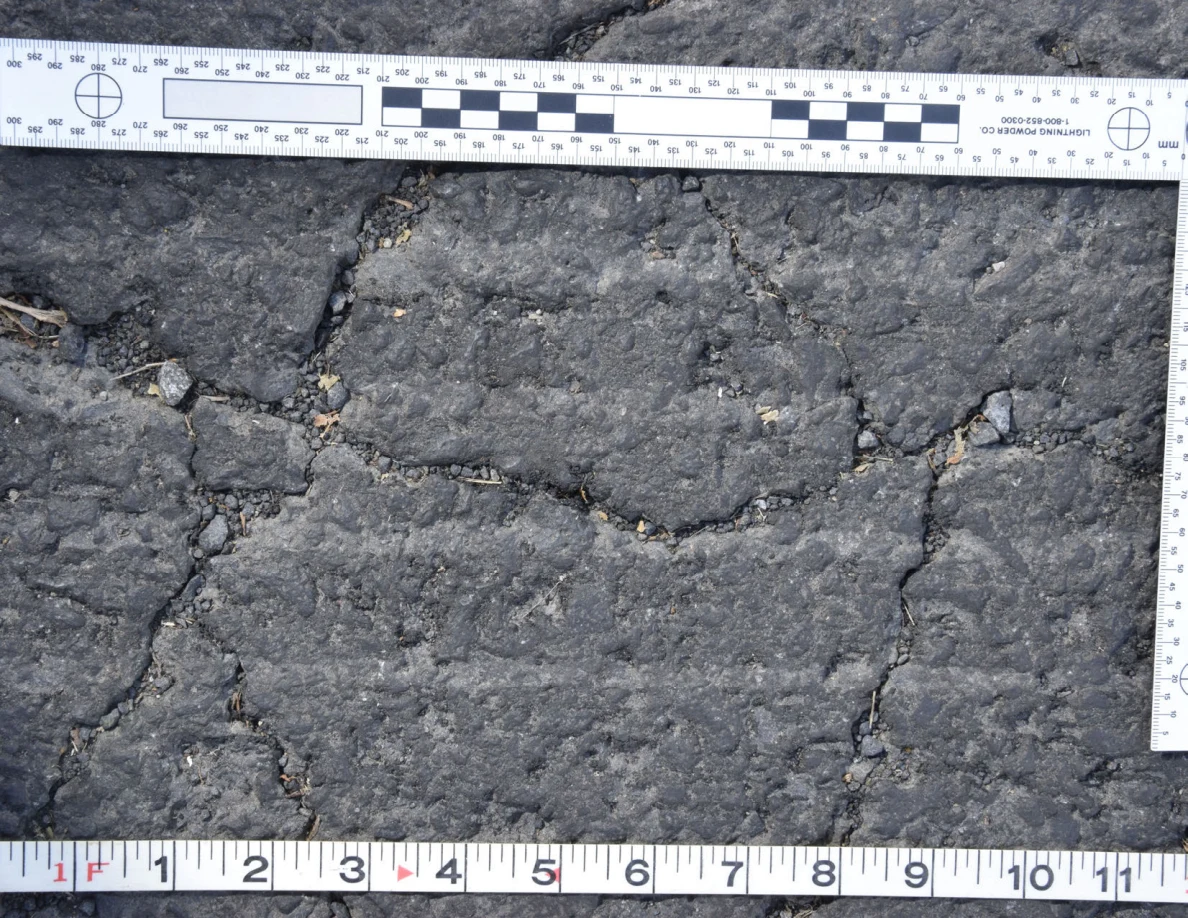 Intersecting cracks in asphalt with small pebbles lodged in the gaps, beside an L-shaped scale and measuring tape.