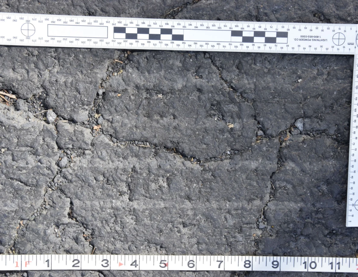 Intersecting cracks in asphalt with small pebbles lodged in the gaps, beside an L-shaped scale and measuring tape.