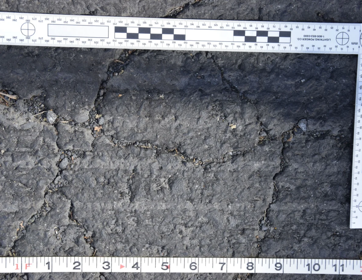 Intersecting cracks in asphalt with small pebbles lodged in the gaps, beside an L-shaped scale and measuring tape.