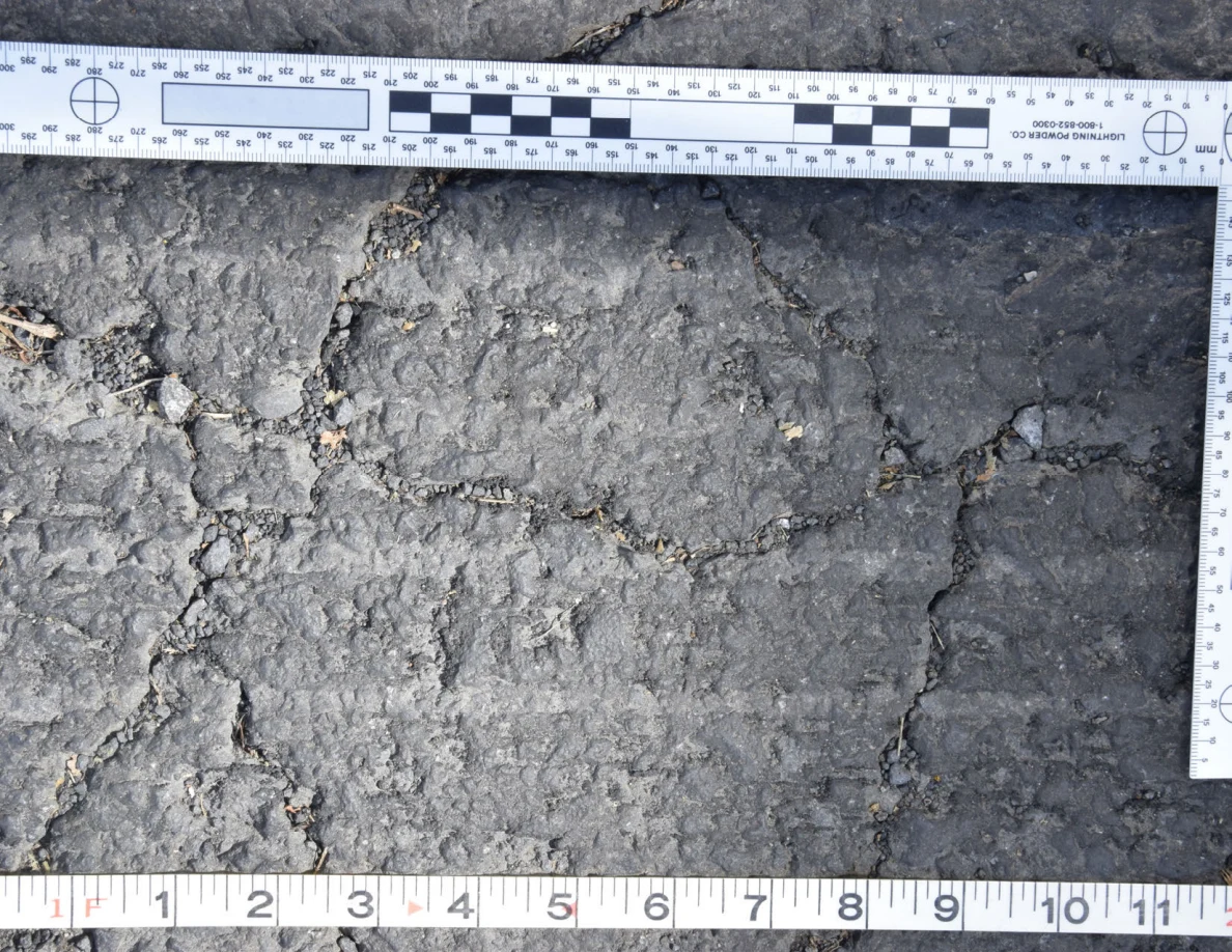 Intersecting cracks in asphalt with small pebbles lodged in the gaps, beside an L-shaped scale and measuring tape.