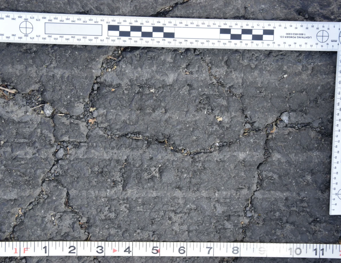 Intersecting cracks in asphalt with small pebbles lodged in the gaps, beside an L-shaped scale and measuring tape.