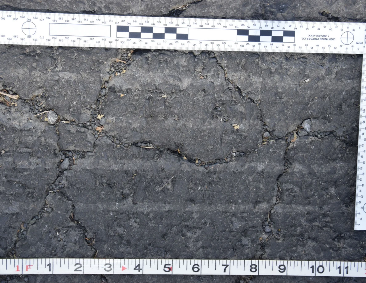 Intersecting cracks in asphalt with small pebbles lodged in the gaps, beside an L-shaped scale and measuring tape.