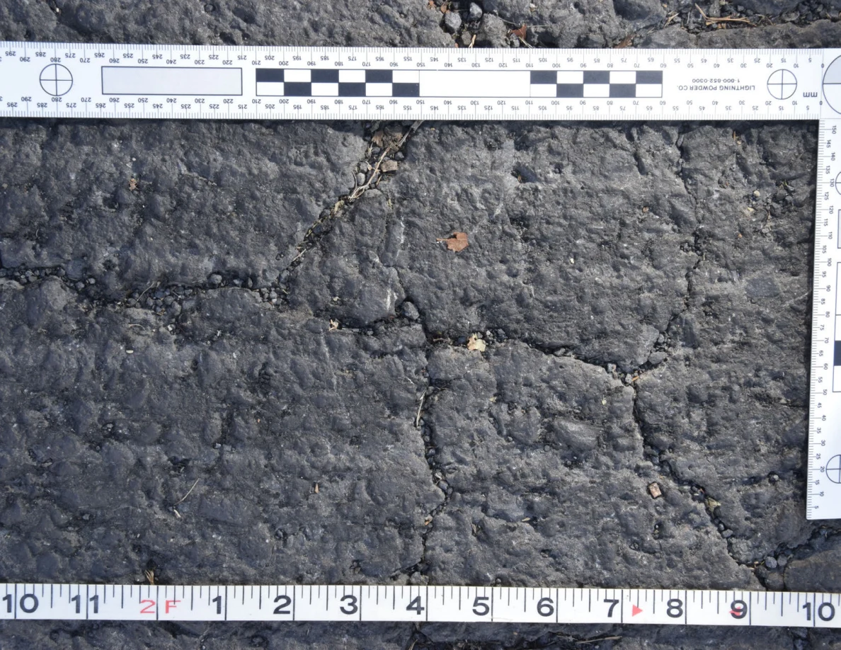 Cracked asphalt with a small brown leaf and grit in the fissures, framed by an L-shaped scale and measuring tape.