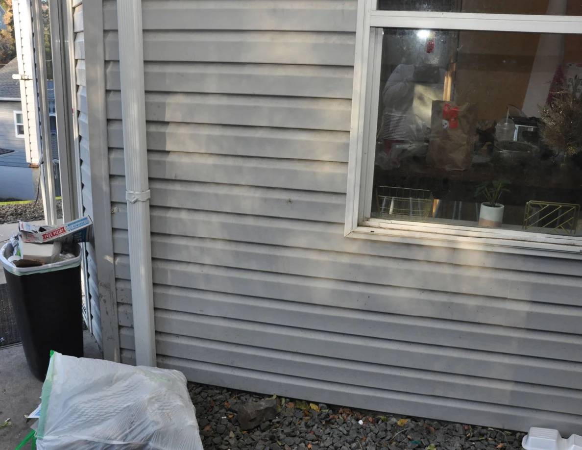 Gray exterior siding, a white-framed window, downspout, trash bin and a white bag on gravel.