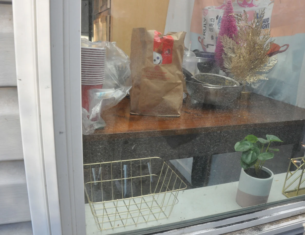 View through a window of a table with paper bags, stacked cups, a metal bowl and small decorative trees; a potted plant and wire baskets sit on the sill.