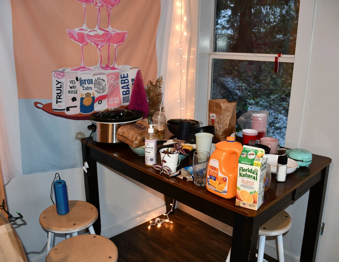 Kitchen table crowded with mugs, cups, drink containers and a slow cooker beneath a colorful drinks-themed wall hanging.