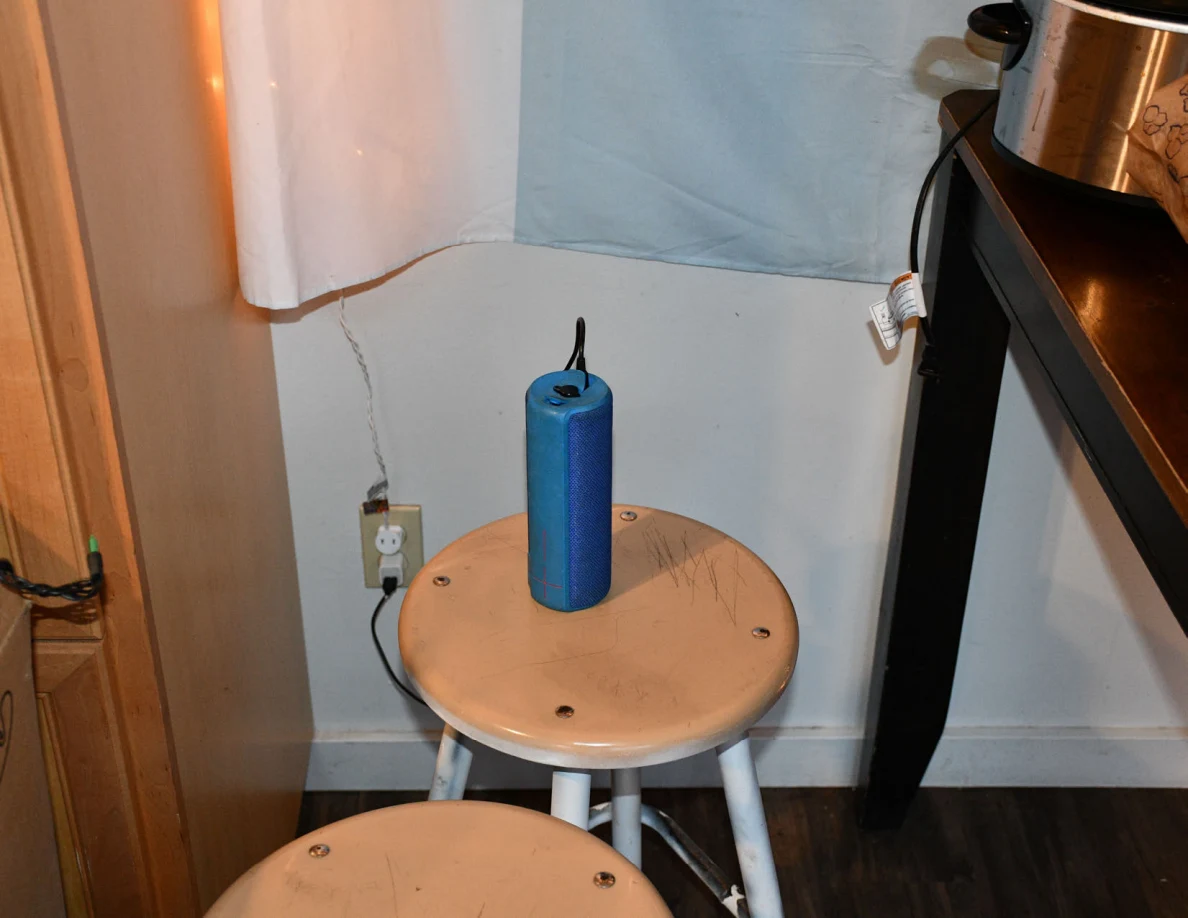 Blue cylindrical portable speaker standing on a wooden stool beside a kitchen table and wall outlet.