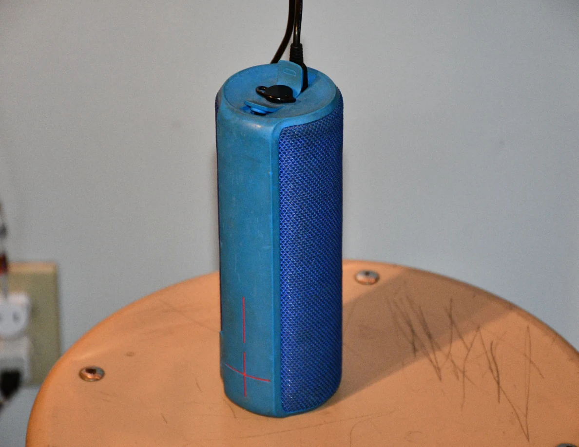 Close view of a blue cylindrical speaker with a black cord on a scratched wooden stool.