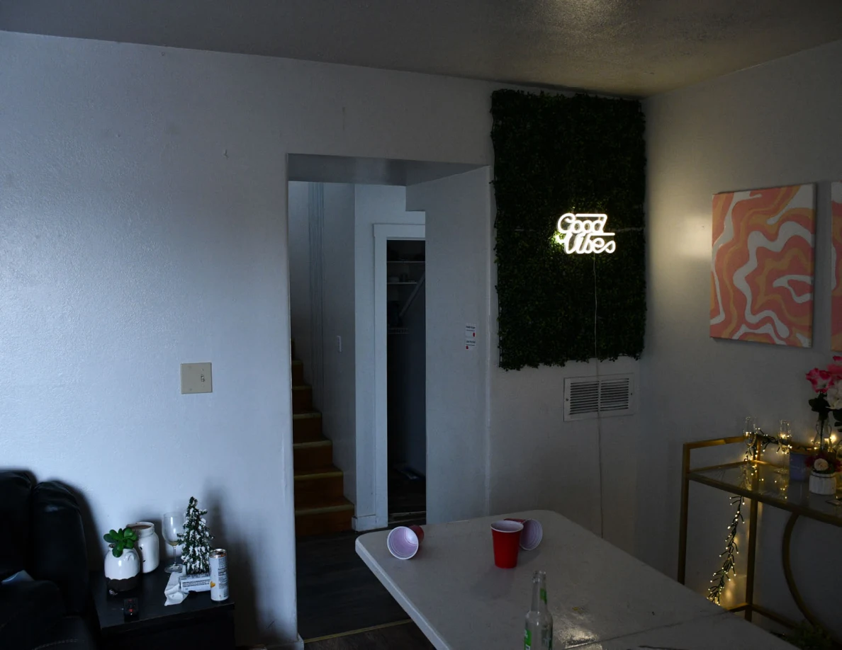 Living room with a white folding table and plastic cups beside a green wall panel with an illuminated Good Vibes sign.