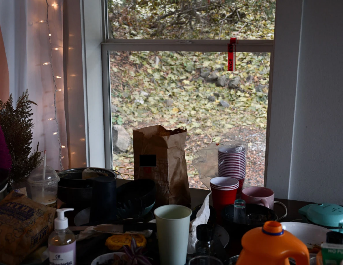 Crowded tabletop below a window, holding stacked cups, mugs, a paper bag, soap bottle and orange drink jug.