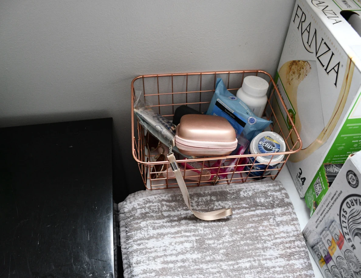 Overhead view of a wire basket holding a pink case and small containers next to boxed drinks.