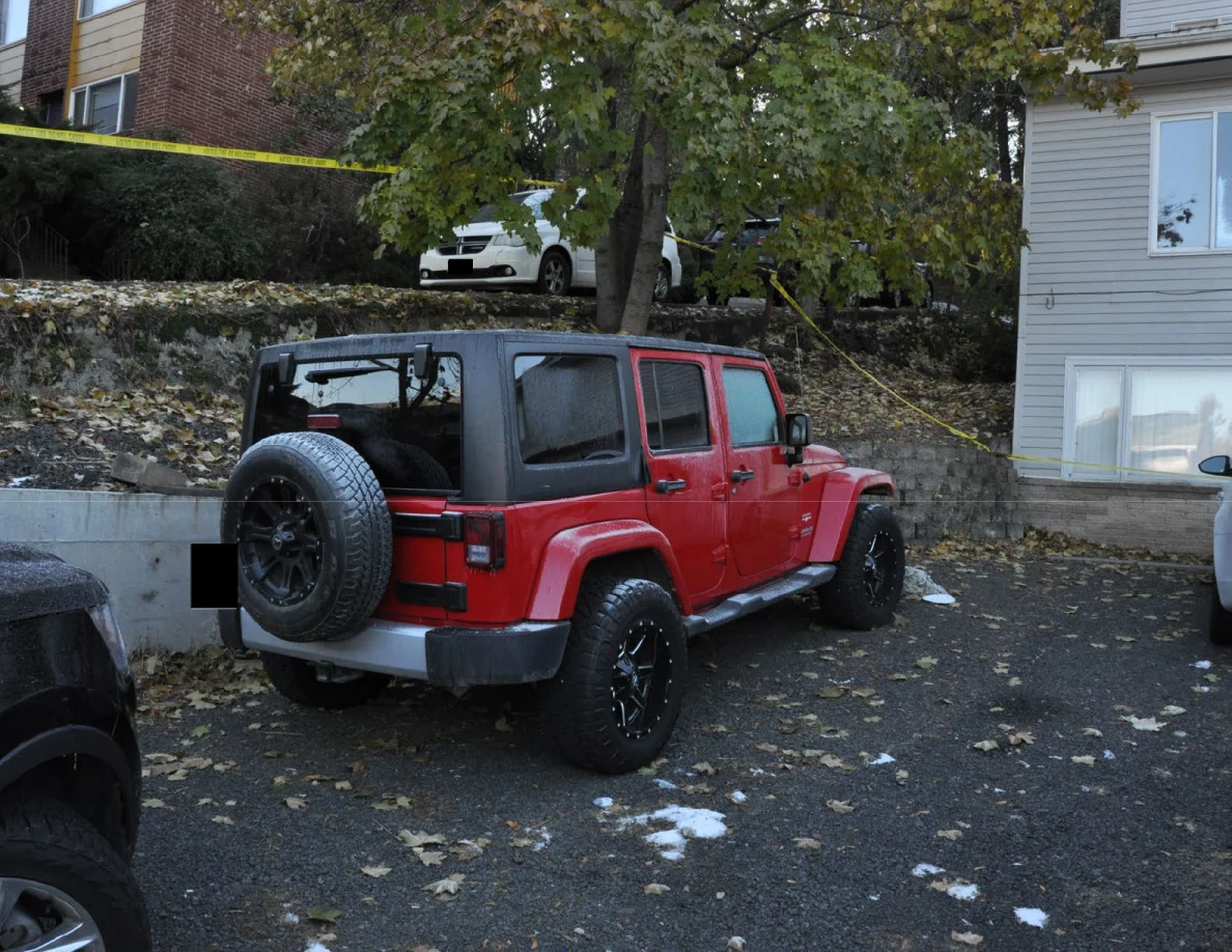 Red Jeep with a rear spare tire parked below a tree and stone retaining wall, with yellow tape nearby.