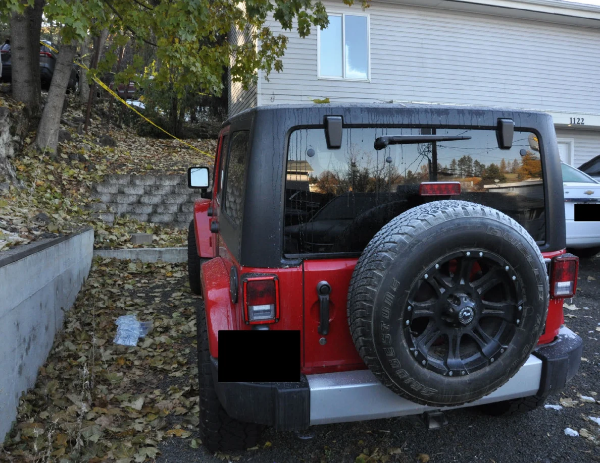 Rear view of a red Jeep with a mounted spare tire beside a house and retaining wall.