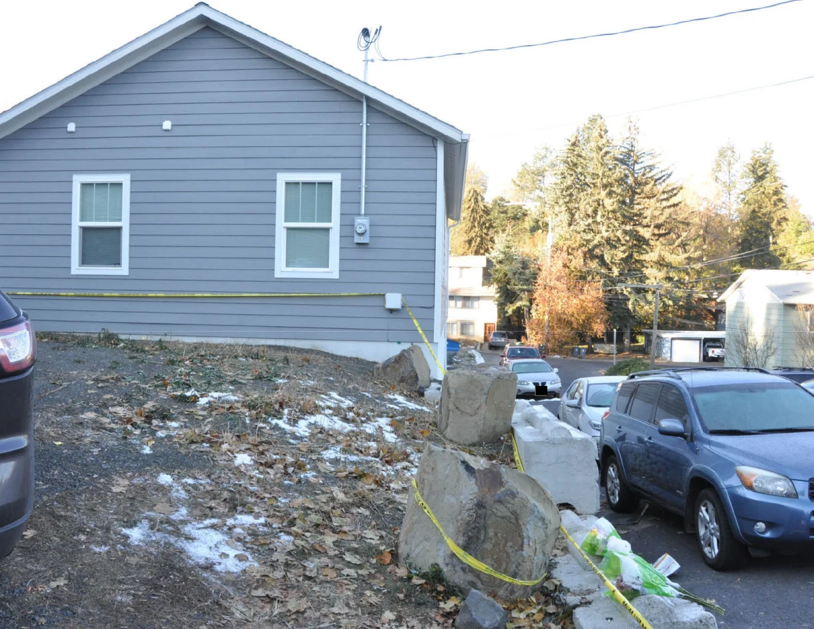 Side of a gray house beside a leaf-covered slope, yellow tape and parked cars.