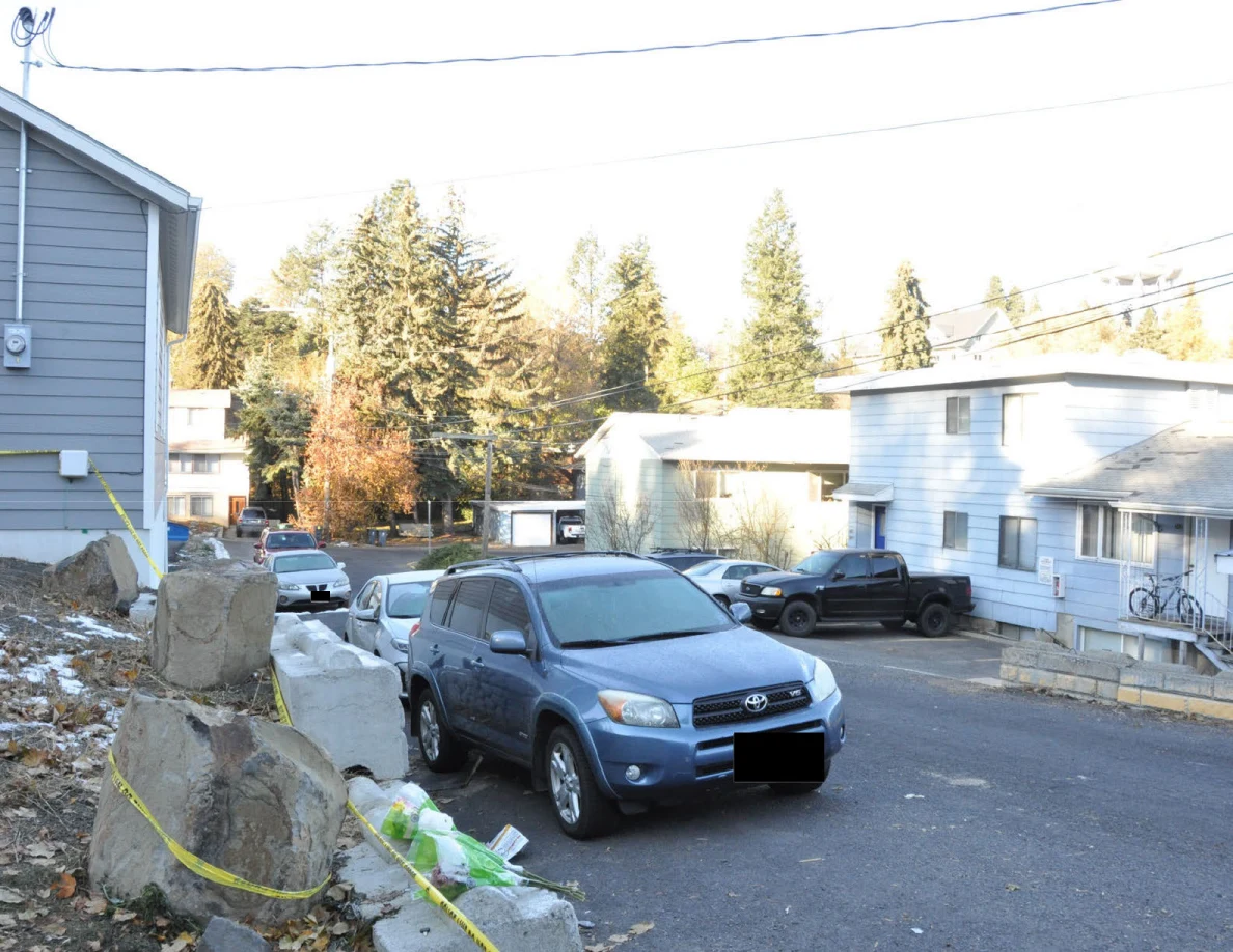 View down a residential street with a blue SUV, stone retaining wall and neighboring buildings.