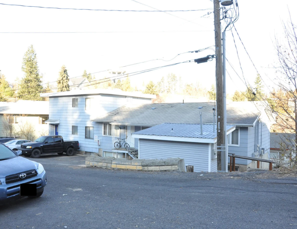 Residential street with light-colored buildings, parked vehicles, a utility pole and overhead wires.