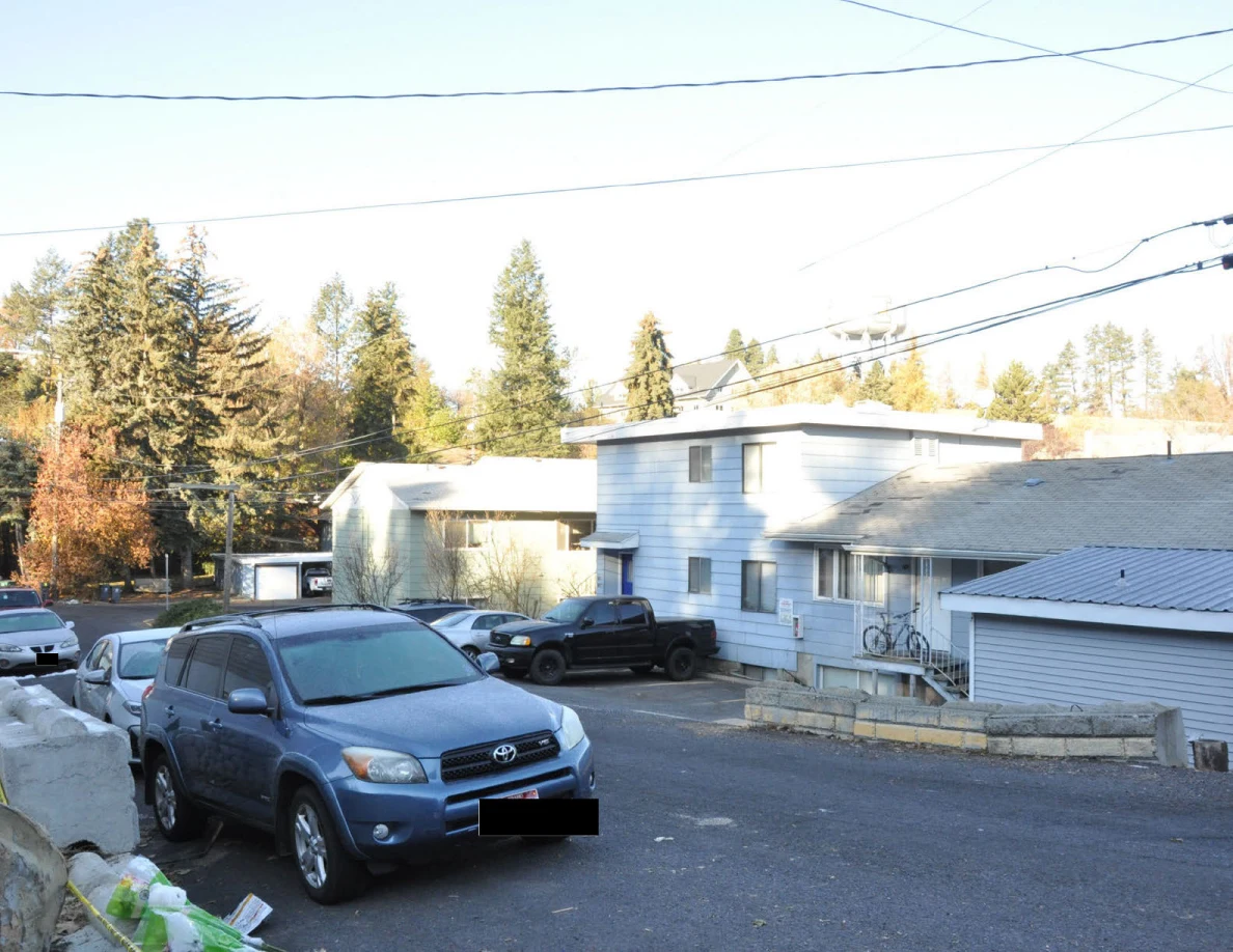 Blue SUV and other parked vehicles on a residential street opposite light-colored buildings.