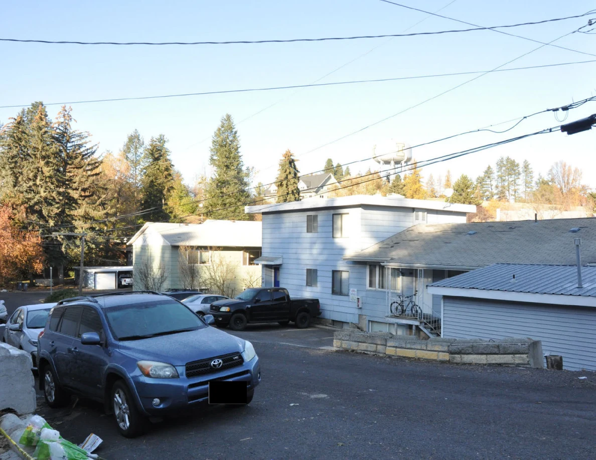Residential street with parked vehicles, light-colored buildings, trees and utility wires.