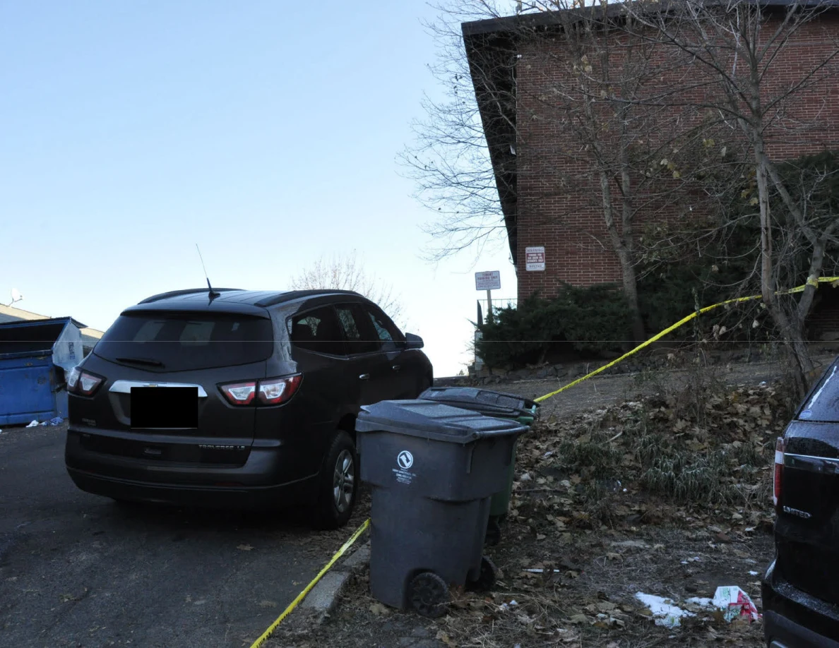 Black SUV and wheeled trash bin beside yellow tape below a brick building.