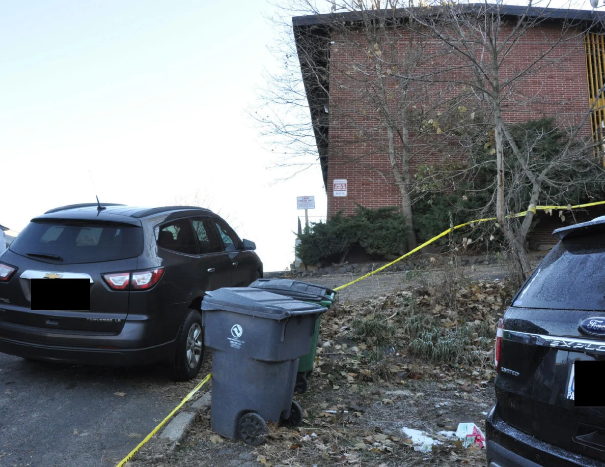 Black SUV and trash bin beside a leaf-covered slope, yellow tape and brick building.