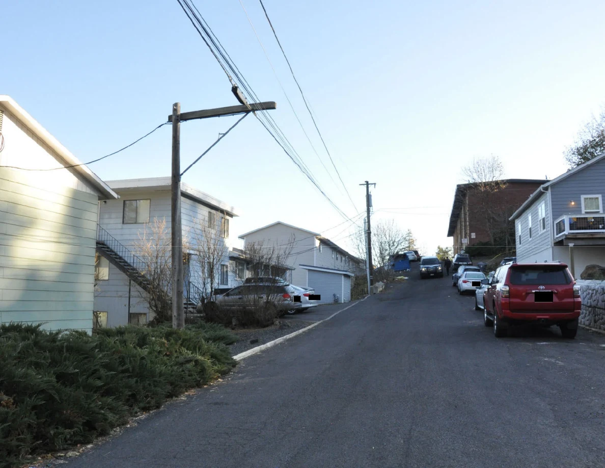 Uphill residential street lined with houses, parked vehicles, wooden utility poles and overhead wires.
