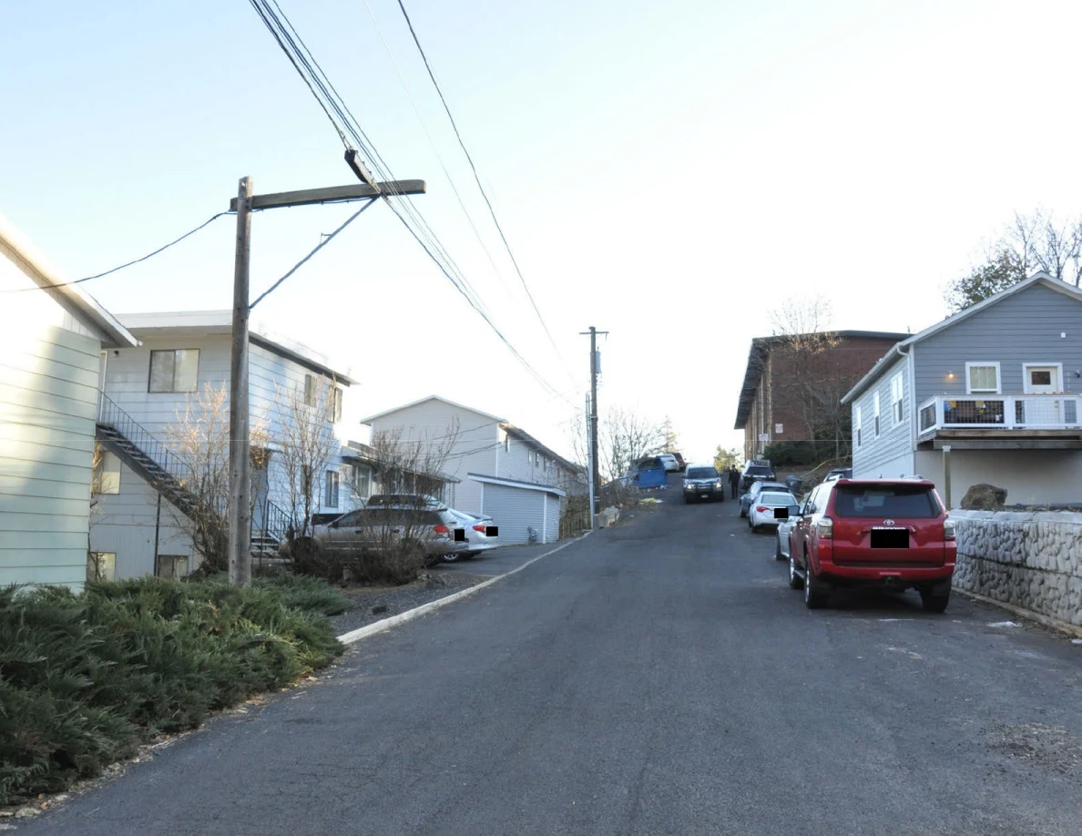 Uphill residential street lined with houses, parked vehicles, wooden utility poles and overhead wires.
