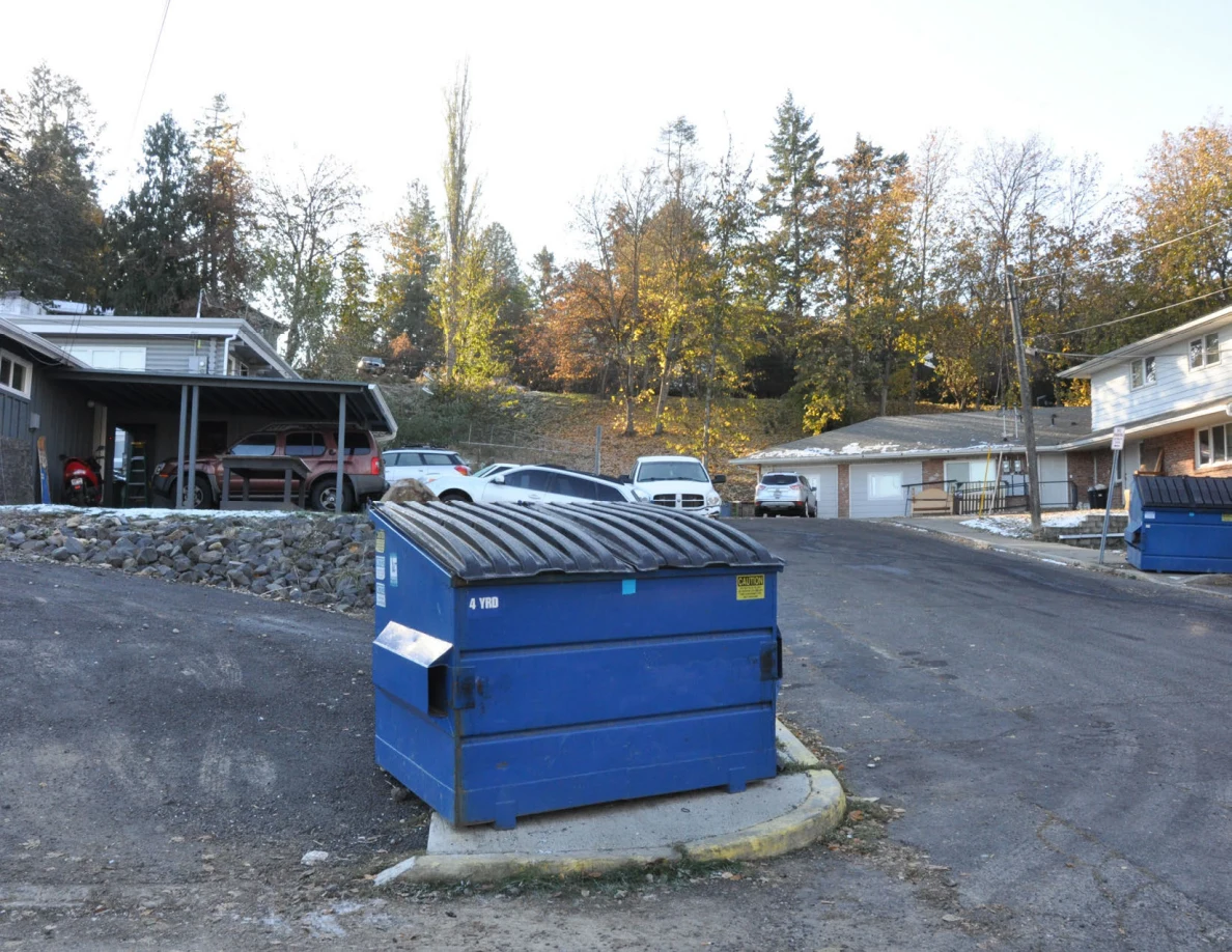 Large blue dumpster beside an asphalt parking area, carports and residential buildings.