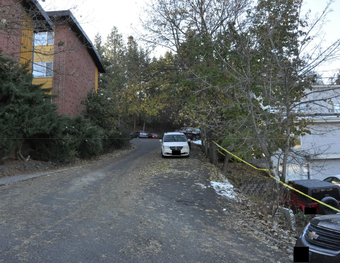 Narrow drive beside a brick apartment building, with parked vehicles, yellow tape and patches of snow.