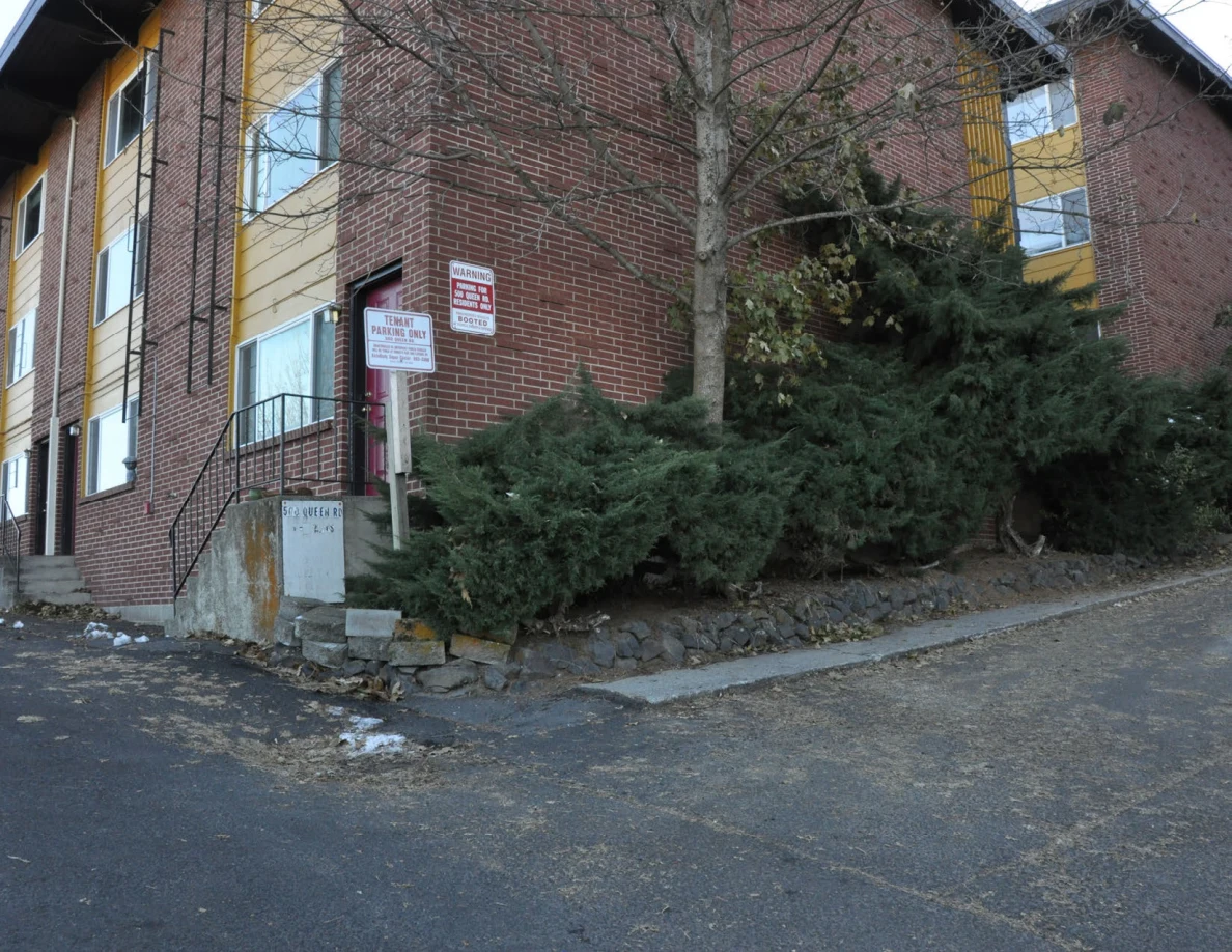 Corner of a brick apartment building with shrubs, entrance steps and posted parking signs.