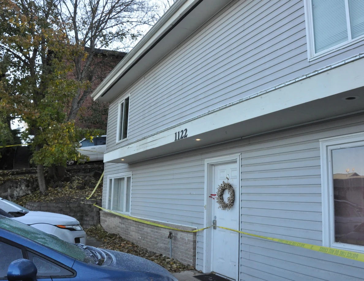 Front of gray-sided house numbered 1122, with a white door, wreath and yellow police tape.