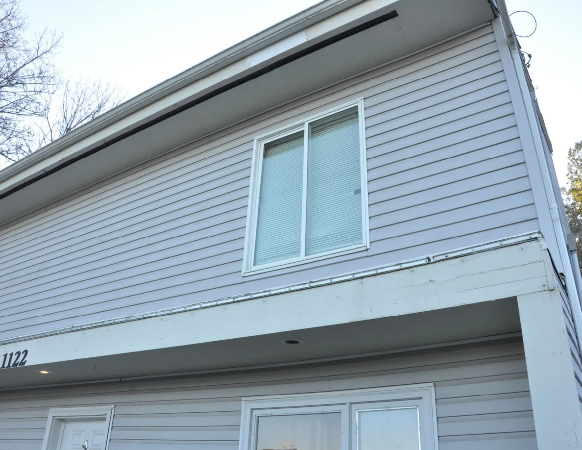 Upward view of a gray-sided house facade, upper windows and roof overhang.