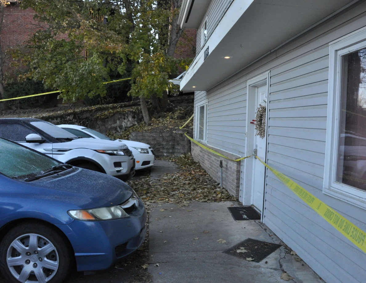 Parked cars beside a gray house entrance crossed by yellow tape, with doormats on the concrete.