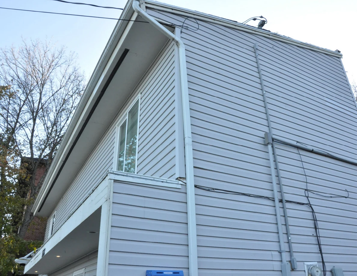 Upper corner of a gray-sided house with roof gutter, downspout and exterior wires.