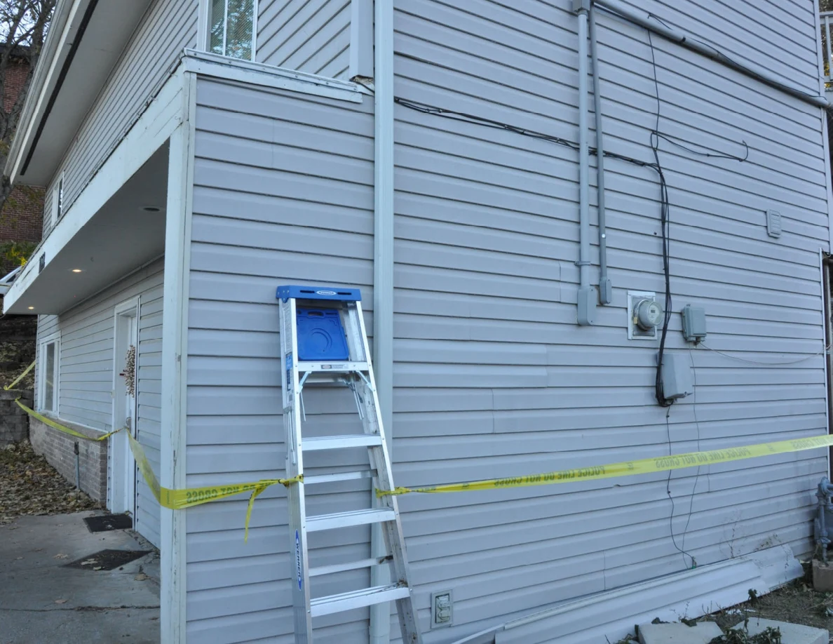 Aluminum ladder leaning against gray siding beside electrical meter equipment and yellow tape.