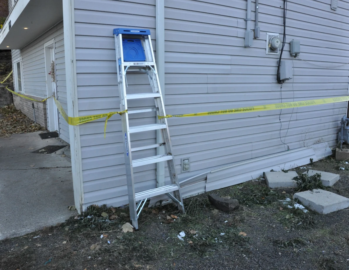 Aluminum ladder at a house corner beside electrical conduits, yellow tape and loose concrete blocks.