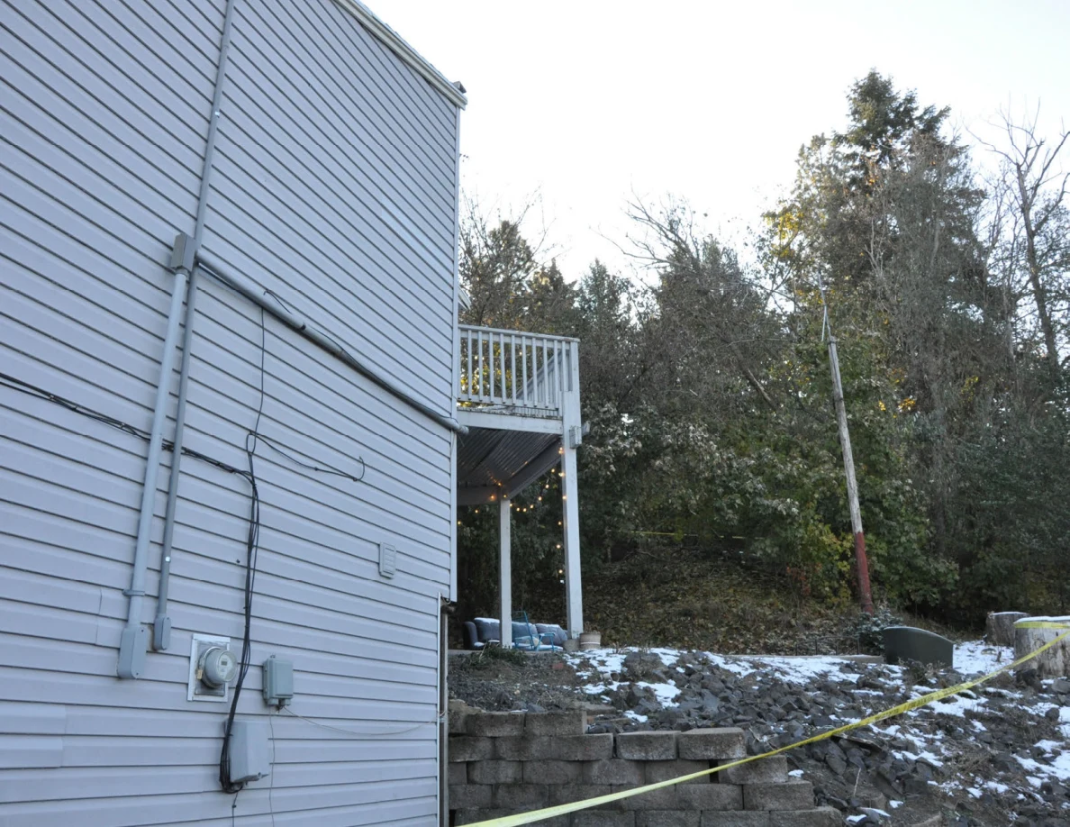 Gray house side wall and raised deck above a rocky slope with snow and a retaining wall.