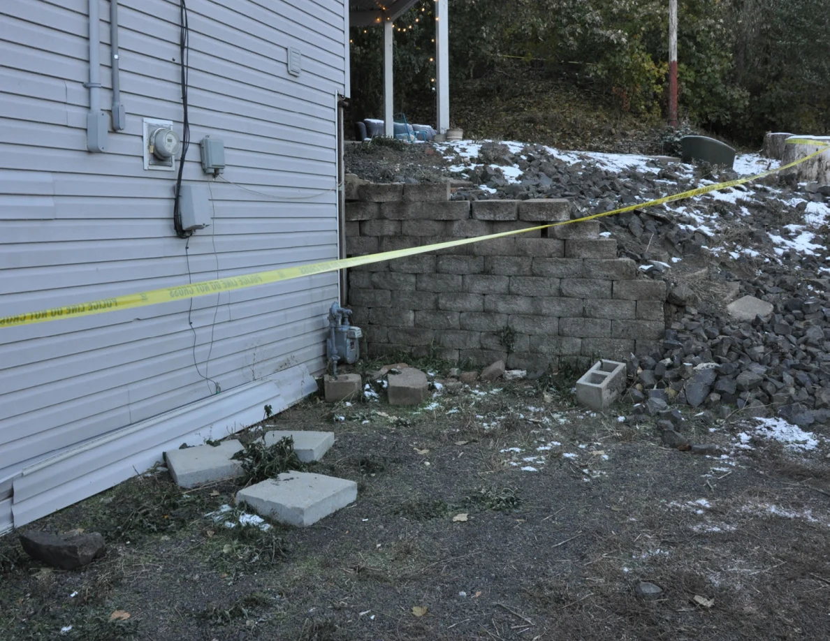 Block retaining wall and loose rocks beside a gray-sided house, with yellow tape across the area.